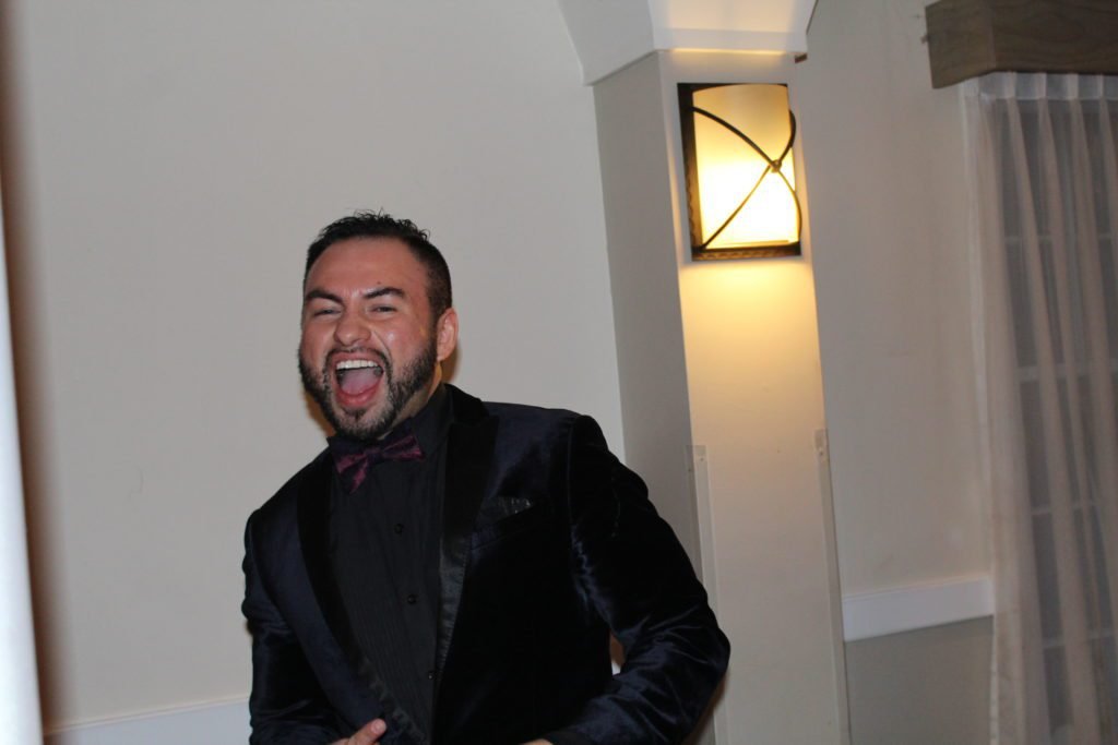 DJ Casanova, a renowned wedding DJ, in a velvet suit with a black shirt and red wine bow tie, joyfully MC'ing at a Latin wedding.