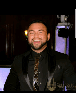 DJ Casanova in a sleek black suite and golden tie, smiling confidently at the camera at a wedding event
